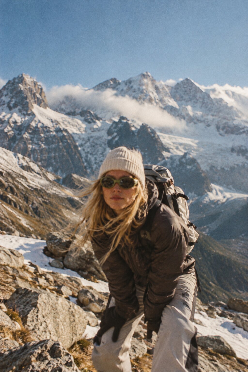 HIKING CLUB: Wanderin mit Mütze und grüner Sonnenbrille vor schneebedeckten Gipfeln